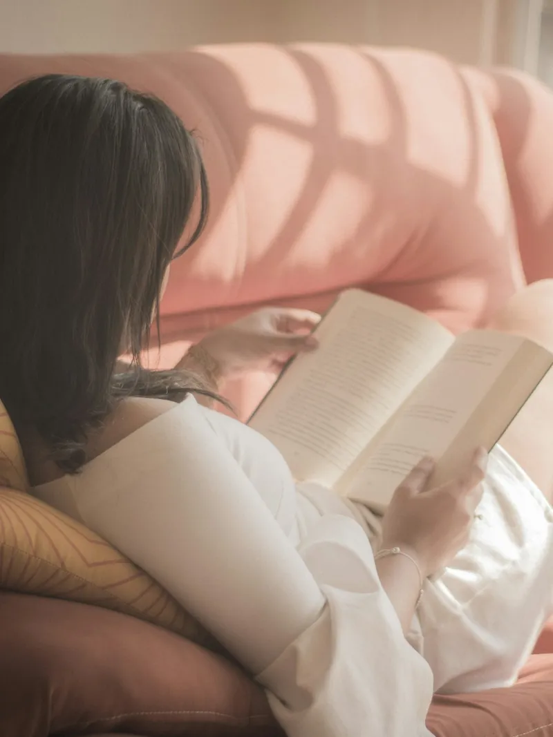 Mujer leyendo un libro recostada en un sillón rosa con la luz de la tarde